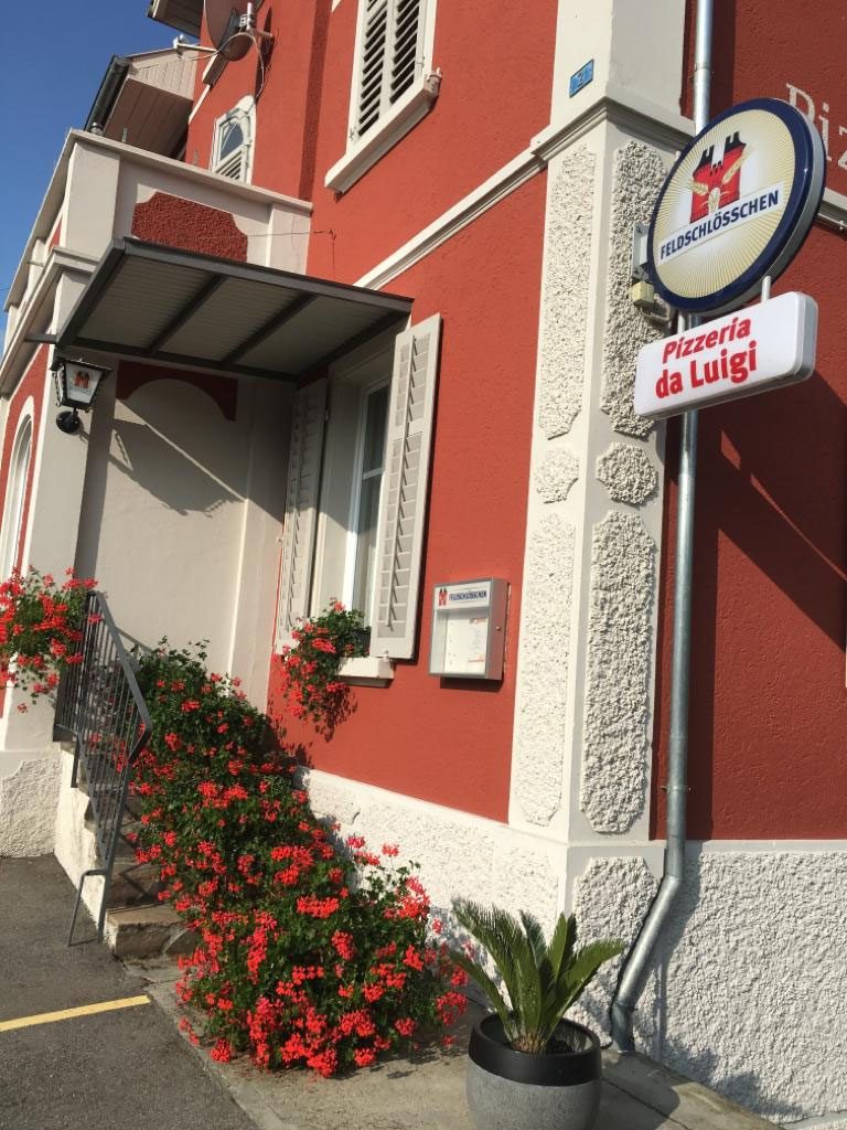 Das Schild Pizzeria da Luigi an der Fassade, darunter blühende Geranien